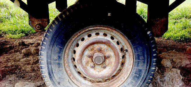 Mittig im Bild der platte Reifen eines Bauwagenrads unter rostiger, eiserner Achskonstruktion und Verbretterung des Aufbaus mit abblätternder, gelber Farbe.