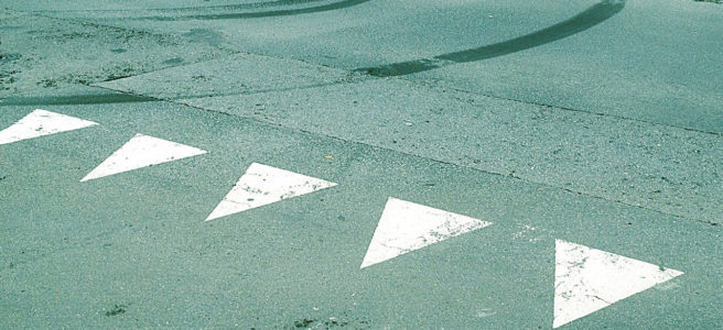 Eine grünlich-bläulich verfärbte Teerfläche. Die Straßenmarkierungen einer Einmündung zeigt kleine weiße Dreiecke, die wie spitze Zähne in Richtung des Betrachters zeigen. Darüber eine nach links kurvende Spur von Gummiabrieb.