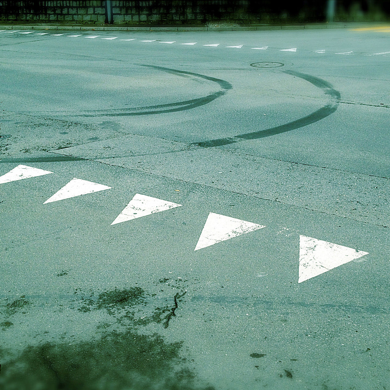 Eine grünlich-bläulich verfärbte Teerfläche. Die Straßenmarkierungen einer Einmündung zeigt kleine weiße Dreiecke, die wie spitze Zähne in Richtung des Betrachters zeigen. Darüber eine nach links kurvende Spur von Gummiabrieb.