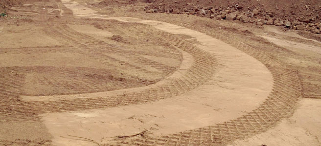 Planiertes Gelände vor einem halb abgerissenen Betongebäude. Die rautenförmige Spur breiter Reifen zeichnen sich im sandigen Untergrund ab und bilden eine 90 Grad Linkskurve.