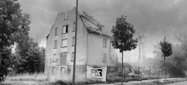 Schwarz-weiß Bild eines Gebäudeabrisses. Die Spitze eines Giebelbaus ist schon zerstört. Im Vordergrund steht eine Straßenlaterne