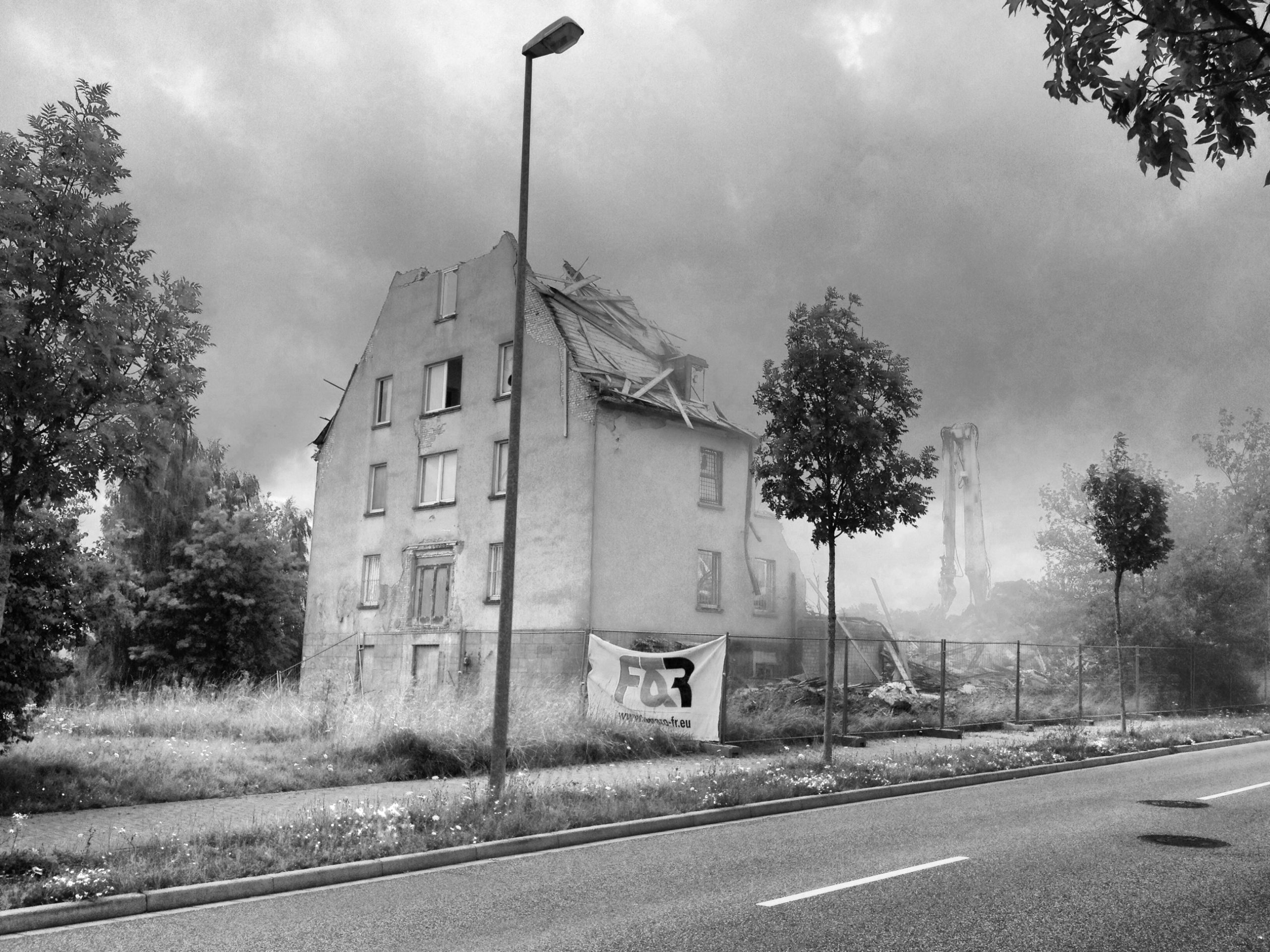 Schwarz-weiß Bild eines Gebäudeabrisses. Die Spitze eines Giebelbaus ist schon zerstört. Im Vordergrund steht eine Straßenlaterne