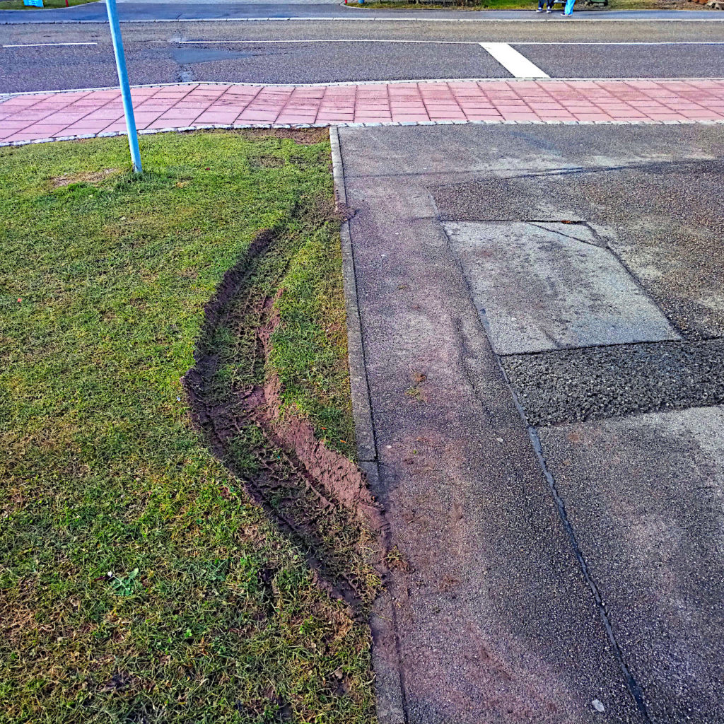 Blick auf eine Straße mit verschiedenen Belägen. Rechts ist Teer, links daneben ein Grünstreifen, in dem eine Spur eines Reifens einen Bogen furcht. Am oberen Bildrand wird die Fläche von roten Pflastersteinen gesäumt.